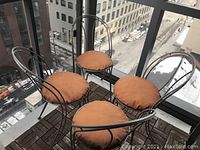 Photo of four matching metal chairs with rounded backs and orange cushions set on a balcony patio with city buildings visible in the background.