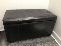 Photo shows a dark brown rectangular bench with leather-like upholstery, visible stitched panels, and a storage lid, standing on small black feet.