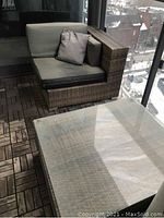 Photo of an outdoor wicker loveseat with green-grey cushions and two matching throw pillows, and part of a glass-top wicker coffee table on patio decking