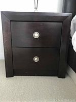 Photo of dark brown wooden side table with two drawers, each drawer has a silver round knob. The table has squared edges and measures approximately 23 inches wide, 16 inches deep, and 24 inches tall.