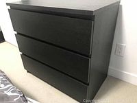 Black-brown IKEA Malm 3-drawer chest showing front and right side on a light carpeted floor next to white baseboard and electrical outlet.