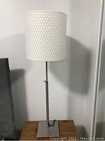 Image of Ikea table lamp with white woven pattern cylindrical shade and metal base on wooden table.
