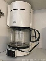 White Braun coffee maker with glass carafe shown on shelf, power cord coiled on front.