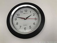 Photo showing a round wall clock with black frame and white face marked with black numbers. The brand 'sky' and 'QUARTZ' are visible.