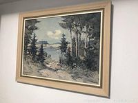 Photo of framed painting depicting a lake and forest landscape with trees, water, and distant hills.
