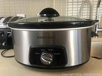 Front view of Black & Decker slow cooker showing stainless steel body, black trim, glass lid with handle, and control dial with Low, High, Warm settings.