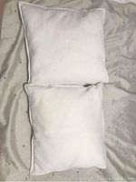 Two small white square pillows lying flat on a light colored surface, showing front view and side seams with piping edge detail.