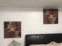 Photo showing two abstract paintings hung on a white wall above a black bed headboard and a lamp nearby.