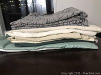 Stack of nine folded queen size flat sheets in assorted colors including grey patterned, white, beige, cream, and green.