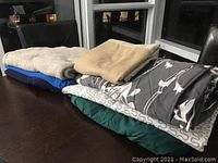 Three fleece blankets, fitted sheet, duvet covers, and queen bed skirt folded and stacked on a dark surface near windows.