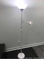 Full view of the white floor lamp showing the pole, round base, white glass upward-facing shade and attached adjustable reading light, plugged into wall outlet.
