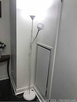 White floor lamp positioned in a corner with the primary light turned on. Visible circular base and upward-facing white conical shade. Secondary smaller reading light arm visible.