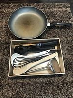Photo showing a black nonstick frying pan and a cardboard box containing multiple cooking utensils including spatulas, whisk, and ladle.