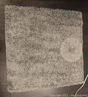 Top-down view showing the full square grey shaggy rug with fringed edges and mottled color tone on dark hardwood floor.
