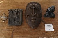 Front view of all four items laid out on a wooden surface: metal spinning top toy, wooden thumb piano, African wooden mask, and black stone carving sculpture.