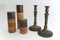 Photo showing two tall ceramic and wicker votive candle holders with tea lights beside two solid brass candlesticks with ornate detailing and round bases.
