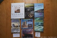 Six books arranged on a wooden surface, showing front covers from left to right: 'The Price Paid for Charley', 'Newfoundland Lighthouses of Atlantic Canada', 'The Frayed Edge', 'God Guard Thee', 'Fort Amherst', and 'An Illustrated Viking Voyage'