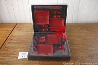 Two unopened Asian servingware sets on a wooden table, showing the contents arranged in black plastic trays within clear plastic packaging.