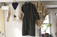 Three items hanging on wall rack: alpaca poncho, green coat with scarf, Newfoundland dog sweater.