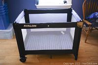 Front view of black Avalon playpen with mesh sides and cushioned base showing geometric patterned fabric, two wheels on one side, metal corner protectors.