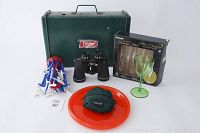 Lot arranged showing green Coleman metal case with stove, binoculars, laundry hanger, orange plates, boxed champagne flutes, green plastic wine glass, mosquito hat cover pouch.