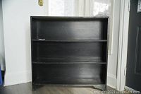 Photo showing front view of black painted shallow bookshelf with three shelves against wall and window.