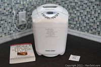 Full view of the breadmaker with the recipe book and instruction manual placed beside it on a countertop, showing overall condition and included items.