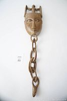 Full view of the wooden mask with a human figure on top and double ringed chain hanging down.
