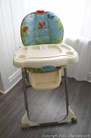 Front view of Fisher Price high chair with padded nature-themed cushion, beige tray with cup holders, and metal legs with wheels.