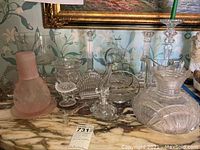 Full lot view showing varied glassware items including candle holders, candy dishes, a pitcher, and a pink frosted perfume dauber on a marble surface.