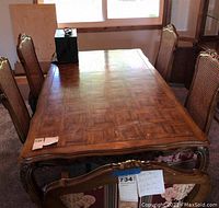 Full view of wooden dining table with carved legs and several woven backed chairs around it, placed near a window. Chairs are not included.