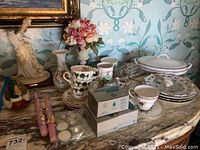 Overview of the lot on a marble table showing the signed porcelain B. Merli statuette, stacked tea cups and saucers, large serving tureen with lid, stacks of decorative plates, pink taper candles, white candles in packaging, floral arrangement in ceramic vase, and a ceramic bird figurine.