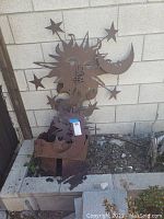 Rusty metal yard art sun and moon attached to metal planter box and separate sun and moon stake in dirt spot by brick wall.