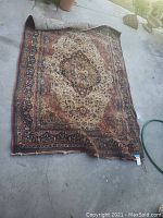Full view of carpet rug laid out on concrete showing central medallion design with folded corner revealing backing and wear.