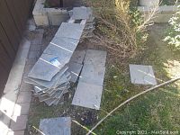 Stack of gray slate tiles laid vertically against a fence, with some tiles on the ground near a garden bed.
