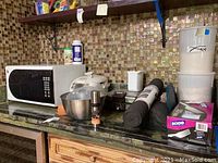 Overview photo of kitchen counter displaying all the lot items including microwave, rice cooker, waffle maker, liners, forks, bowl, containers, and grinder.