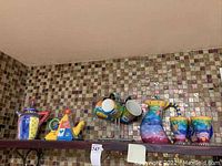 Photo showing a multicolor ceramic pitcher, colorful teapot, and four mugs hanging on a rotating stand against a tiled backsplash.