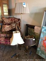 Photo showing three lamps: tall floor lamp with pleated beige shade, mid-sized lamp with white shade, and shorter lamp with detached brown shade. Lamps placed near patterned armchair and paintings.