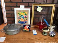 Photo shows a variety of decorative items on a wooden surface against a brick wall. Items include a metal tortilla server, small framed artwork, gold picture frame, two glass bird paperweights, a red glass art vase, and a colorful handmade artwork.