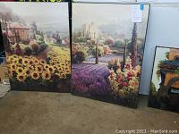 Two larger paintings with countryside flower fields and buildings, resting on floor leaning against wall.