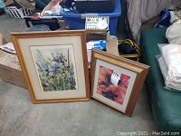 Two framed floral art prints placed on a floor beside storage boxes and other items.