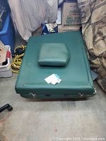 Top-down view of green massage table folded flat with attached padded armrest, showing worn vinyl surface and metal latches.