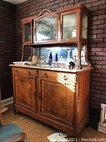 Front and slightly angled view of vintage wooden display cabinet showing granite top, two glass upper doors, and wooden lower doors with brass hardware, placed against brick wall.