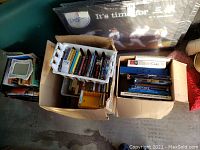 Two cardboard boxes, one with assorted cassette tapes loosely arranged, the other with various books mostly hardcover and some paperback related to vehicles and geography.