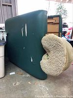 Side view of teal massage table showing several tears in leather upholstery and the separate padded crescent face cradle with shearling cover placed next to it.