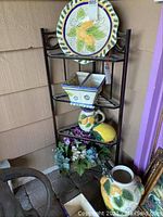 Black metal corner rack with four shelves shown with decorative plates, ceramic containers, and artificial flowers placed on shelves to illustrate size, but contents not included.