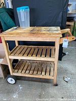 Full view of wooden kitchen cart showing two shelves, handles, and wheels. Surface shows discoloration and signs of wear.