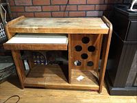 Front view of oak wine cart showing top surface, white sliding drawer, open lower shelf, and vertical bottle holder section with circular holes.