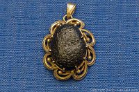 Front view of pendant showing oval meteorite fragment in decorative gold-tone metal frame.