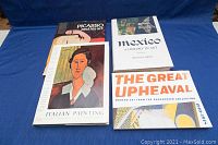Four art books laid flat on blue cloth background: 'Picasso and His Art', 'Mexico: A History in Art', 'Italian Painting: From Caravaggio to Canaletto', 'The Great Upheaval'.